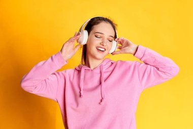 Pembe kapüşonlu güzel bir kızın portresi sarı stüdyo arka planında kulaklıkla müzik dinliyor. Gençlik kavramı, güzellik, moda, yaşam tarzı, duygular, yüz ifadesi. Ad