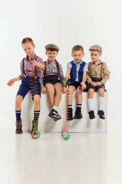 Bir grup küçük çocuk, klasik retro giysili çocuklar gri stüdyo arka planında oturup oyuncaklarla oynuyorlar. Oyun konsepti, çocukluk, arkadaşlık, etkinlik, boş zaman, eski tarz, moda