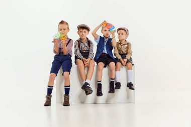 Bir grup küçük çocuk, klasik retro giysili çocuklar gri stüdyo arka planında oturup oyuncaklarla oynuyorlar. Oyun konsepti, çocukluk, arkadaşlık, etkinlik, boş zaman, eski tarz, moda