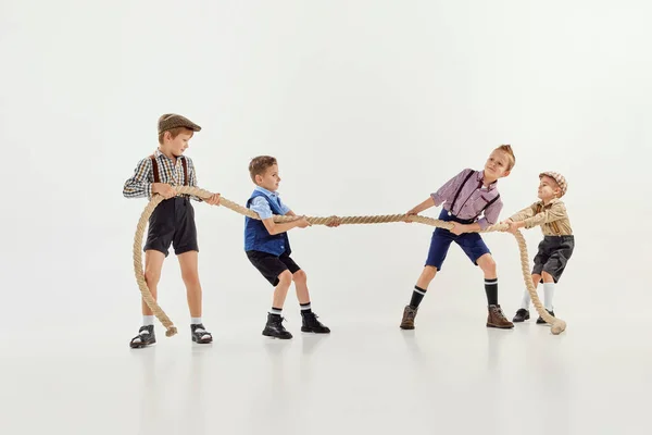 Competition. Group of little boys, children playing together, pulling the rope over grey studio background. Concept of game, childhood, friendship, activity, leisure time, retro style, fashion.