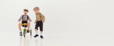 Çocuklar, gri stüdyo arka planında antika kıyafetlerle bisiklet süren çocuklar. Pankart, broşür. Oyun konsepti, çocukluk, arkadaşlık, etkinlik, boş zaman, eski tarz, moda.