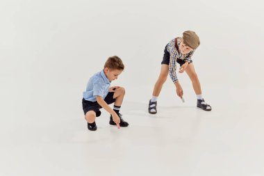 İki çocuk, klasik retro giysili çocuklar yere tebeşirle gri stüdyo arka planlarını çiziyorlar. Oyun konsepti, çocukluk, arkadaşlık, etkinlik, boş zaman, eski tarz, moda.