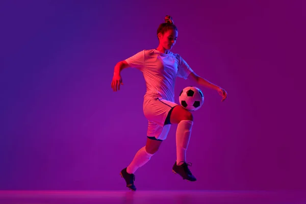 Knee kick. Young professional female football, soccer player in motion, training, playing over gradient pink background in neon light. Concept of sport, action, motion, goals, competition, hobby, ad.