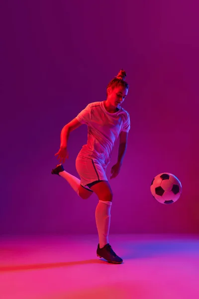 Winner. Young professional female football, soccer player in motion, training, playing over gradient pink background in neon light. Concept of sport, action, motion, goals, competition, hobby, ad.