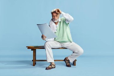 Yakışıklı, ciddi, şık giyinmiş, mavi arka planda spor dergisiyle bankta oturan son sınıf öğrencisinin portresi. Boş zaman aktivitesi kavramı, hobi, yaşam tarzı, fitness, duygular, eski tarz