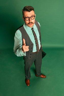 Top view. Handsome man, businessman in classical suit with briefcase and glasses showing gesture of approvement over green background. Concept of emotions, business, occupation, facial expression