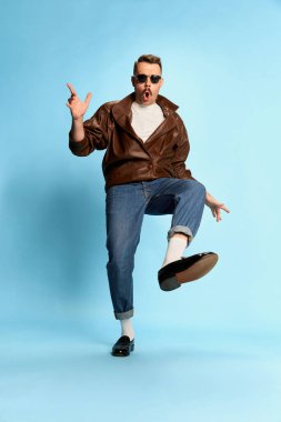 Portrait of brutal, handsome man in jeans, leather jacket and sunglasses posing, dancing over blue studio background. Concept of emotions, facial expression, mens fashion, lifestyle. Ad