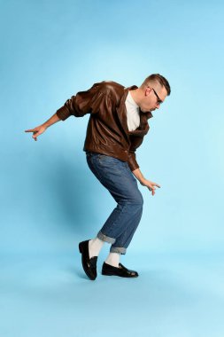 Joy and energy. Portrait of brutal, handsome man in jeans, leather jacket and sunglasses posing, dancing on blue studio background. Concept of emotions, facial expression, mens fashion, lifestyle. Ad