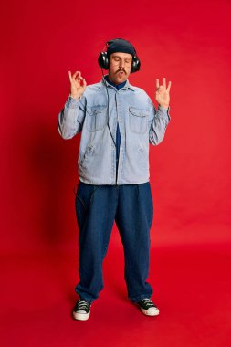 Portrait of man in stylish, oversized, modern clothes, listening to music in headphones over red background. Concept of modern fashion, lifestyle, music culture, emotions, facial expression. Ad