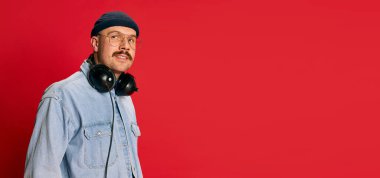 Portrait of man in stylish modern clothes, glasses, hat and headphones posing over red background. Banner, flyer. Concept of modern fashion, lifestyle, music culture, emotions, facial expression. Ad