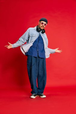 Portrait of man in stylish clothes, oversized jeans, shirt, hat and headphones posing over red background. Concept of modern fashion, lifestyle, music culture, emotions, facial expression. Ad