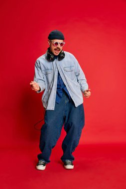 Portrait of man in stylish modern clothes, oversized jeans, shirt and hat posing in headphones over red background. Concept of modern fashion, lifestyle, music culture, emotions, facial expression. Ad