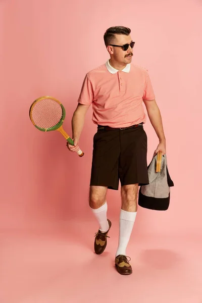 Country sport club member. Portraits of handsome charismatic man in stylish clothes going to play tennis over pink studio background. Concept of sport, emotions, retro style, lifestyle, fashion. Ad