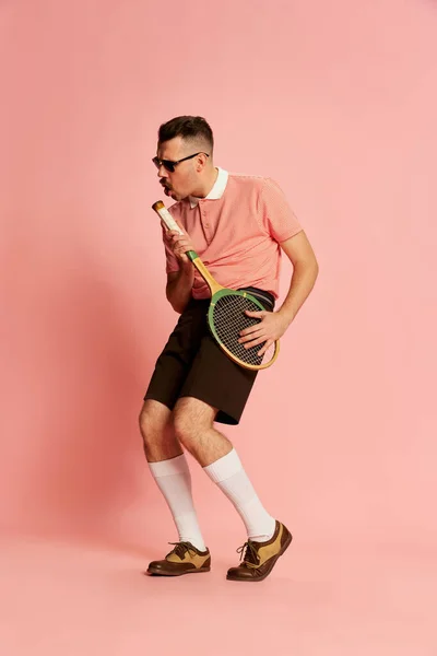 Singer. Portraits of handsome charismatic man in stylish clothes posing with tennis racket over pink studio background. Concept of sport, emotions, retro style, lifestyle, fashion. Ad