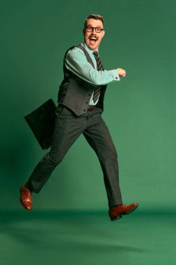 Happy worker. Portrait of handsome man, businessman in classical suit with briefcase over green studio background. Concept of emotions, business, occupation, facial expression, fashion