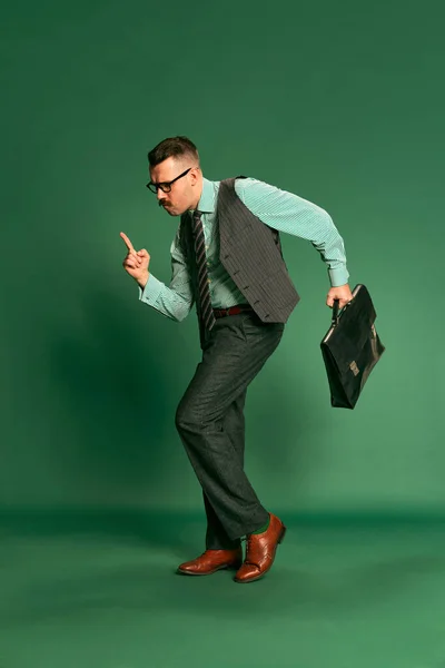 Successful project. Portrait of handsome man, businessman in classical suit with briefcase dancing over green studio background. Concept of emotions, business, occupation, facial expression, fashion