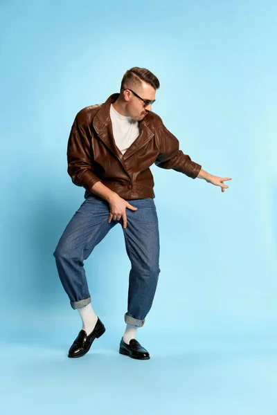 Self-expression. Portrait of brutal, handsome man in jeans, leather jacket and sunglasses posing, dancing over blue background. Concept of emotions, facial expression, mens fashion, lifestyle. Ad