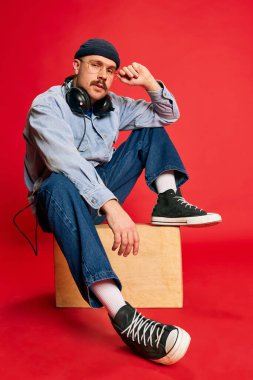 Portrait of man in stylish clothes, oversized jeans, shirt, hat and headphones posing over red background. Concept of modern fashion, lifestyle, music culture, emotions, facial expression. Ad