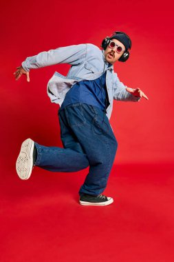 Portrait of man in stylish clothes, oversized jeans, shirt and hat posing, dancing over red background. Concept of modern fashion, lifestyle, music culture, emotions, facial expression. Ad