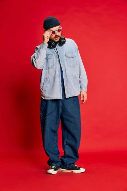 Portrait of man in stylish modern clothes, oversized jeans, shirt and hat posing over red background. Concept of modern fashion, lifestyle, music culture, emotions, facial expression. Ad