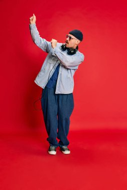 Dance. Portrait of man in stylish modern clothes, oversized jeans, shirt and hat posing over red background. Concept of modern fashion, lifestyle, music culture, emotions, facial expression. Ad