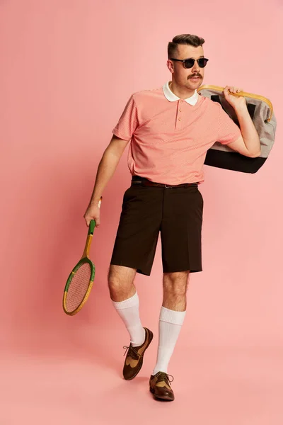 Sport club member. Portraits of handsome charismatic man in stylish clothes posing with tennis racket over pink studio background. Concept of sport, emotions, retro style, lifestyle, fashion. Ad