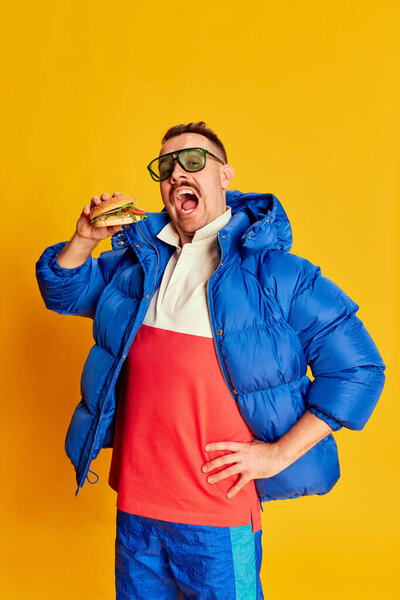 Портрет красивого мужчины в синей зимней куртке, позирующего с бургером на ярко-желтом фоне. Нездоровая еда. Концепция досуга, зимние каникулы, эмоции, спорт, выражение лица, веселье. Объявление