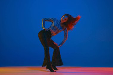 Dynamics. Portrait of young girl dancing high heel dance in stylish clothes over blue background in neon light. Concept of dance lifestyle, modern style, contemporary, youth culture, self-expression