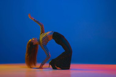 Flexibility. Portrait of young girl dancing high heel dance in stylish clothes on blue background in neon light. Concept of dance lifestyle, modern style, contemporary, youth culture, self-expression