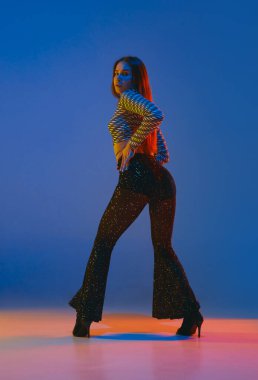 Portrait of young girl dancing heels dance in stylish sparkling clothes over blue background in neon light. Concept of dance lifestyle, modern style, contemporary, youth culture, self-expression