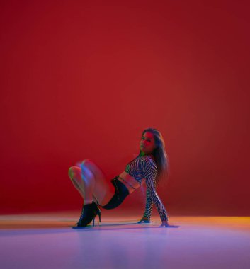 Warming-up. Portrait of young girl dancing heels dance in stylish clothes on red background in neon light. Concept of dance lifestyle, modern style, contemporary dance, youth culture, self-expression