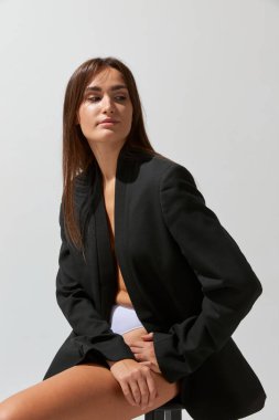 Portrait of young attractive girl posing in black classic suit and underwear over grey studio background. Concept of body and skin care, fitness, natural beauty, health, wellness.
