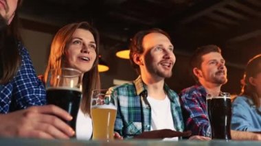 Group of young people, men and women watching match at pub, drinking beer. Fans emotionally cheering up sport team. Celebration of winning game. Competition, championship, match translation. Emotions.
