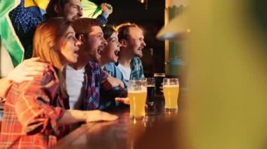 Friends,young people, men and women watching football match at pub. Fans emotionally cheering up Brazil team. Drinking beer. Competition, championship, match translation. Emotions.