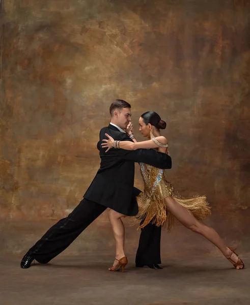 Attraction, passion, emotions. Man and woman, professional dancers in stylish, beautiful stage costumes performing ballroom dance over dark vintage background. Concept of hobby, lifestyle, action