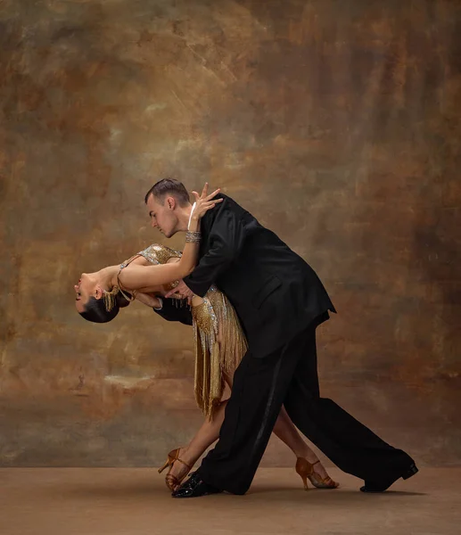 Passionate man and woman, professional dancers in stylish stage costumes performing ballroom, tango dance over dark vintage background. Concept of hobby, lifestyle, action, motion.