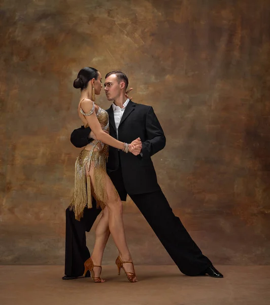 Deep, impressive look. Man and woman, professional tango dancers in stylish, beautiful stage costumes performing over dark vintage background. Concept of hobby, lifestyle, action, motion.