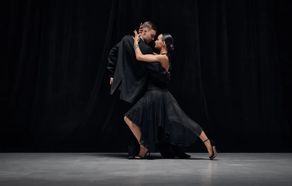 Passionate dance style. Man and woman, professional tango dancers performing in black stage costumes over black background. Concept of hobby, lifestyle, action, motion, art, dance aesthetics