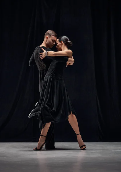 Sensual dance type. Man and woman, professional tango dancers performing in black stage costumes over black background. Concept of hobby, lifestyle, action, motion, art, dance aesthetics