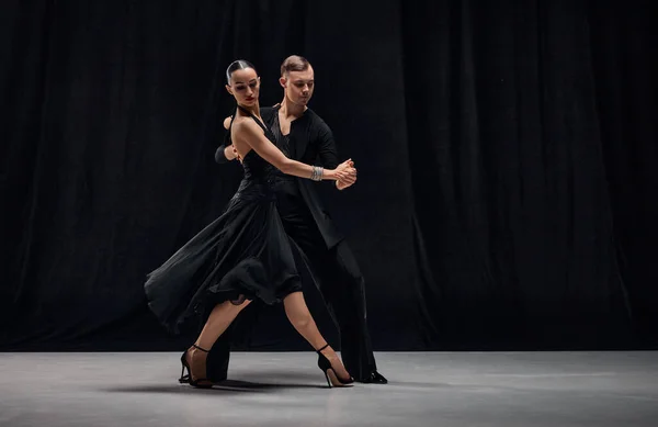 Impressive passionate performance. Man and woman, professional tango dancers performing in black stage costumes on black background. Concept of hobby, lifestyle, action, motion, art, dance aesthetics