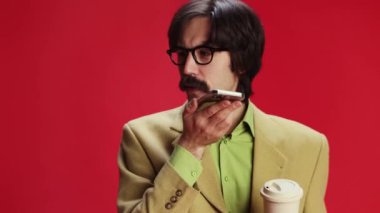 Man, businessman in classic suit recording voice message on phone and drinking coffee over red background. Communication. Concept of emotions, business, occupation, facial expression, fashion