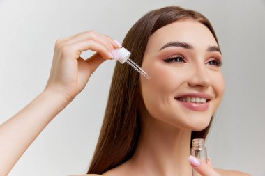 Attractive young girl with well-kept healthy skin applying face serum, posing over grey studio background. Concept of natural beauty, skin care, cosmetology, cosmetics, health, plastic surgery