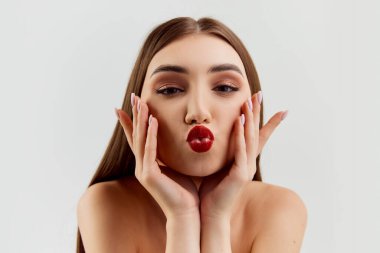 Sending kisses. Young beautiful brunette girl with red lips makeup posing over grey studio background. Tenderness. Concept of natural beauty, youth, fashion, cosmetology, wellness, makeup.