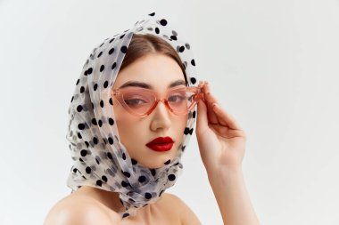 Beautiful young girl wearing trendy sunglasses and scarf on head, posing over grey studio background. Femininity vibe. Concept of natural beauty, youth, fashion, cosmetology, wellness, makeup.