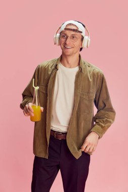 Young positive man in stylish clothes listening to music in headphones, drinking orange juice, posing over pink studio background. Concept of emotions, facial expression, lifestyle, retro fashion. Ad
