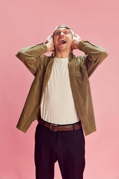 Burst of laugh. Young positive man posing, emotionally listening to music in headphones over pink studio background. Concept of emotions, facial expression, lifestyle, retro fashion. Ad