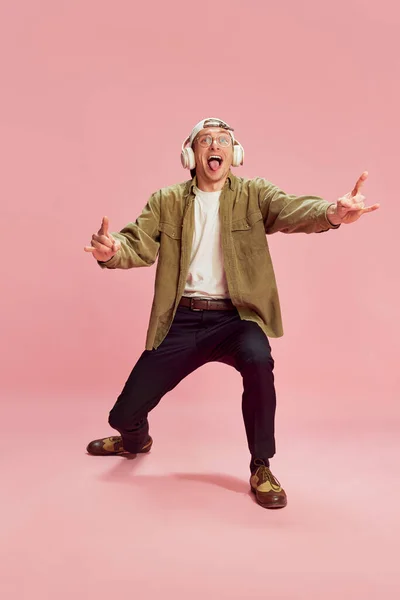 Rock and roll generation. Young positive man posing, emotionally listening to music in headphones over pink studio background. Concept of emotions, facial expression, lifestyle, retro fashion. Ad