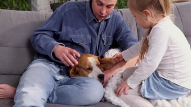Happy family cuddling their dog beagle, having fun together in room. Concept of pets, parenthood, love for animals. Dad and daughter sitting on the sofa at home, playing and relaxing. Friendship