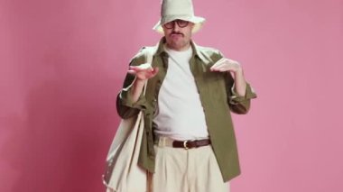 Joyful tourist. Emotive man in panama with big shopper bag, in comfortable clothes dancing, making funny movements over pink studio background. Concept of emotions, lifestyle, mens fashion, mood. Ad