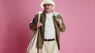 Having fun on vacation. Stylish emotive man, tourist in comfortable clothes dancing with funny movements, grimacing over pink background. Concept of emotions, lifestyle, mens fashion, mood. Ad